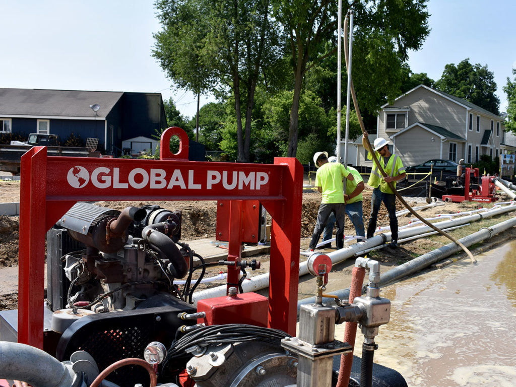 Red GLOBAL PUMP unit with hoses actively dewatering residential construction site—three workers in safety gear managing equipment near wet ground and nearby homes.